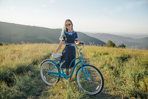 Beautiful young woman in dress standing with bicycle cruiser on rural road along green summer meadows and looking at camera.