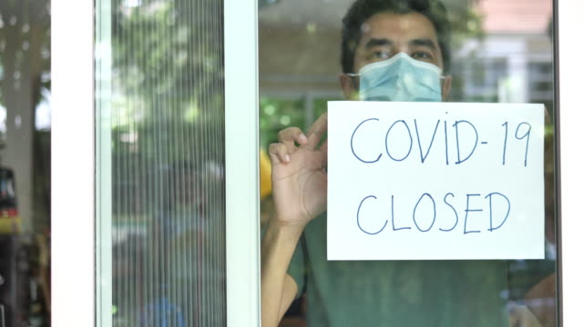 Coronavirus closed for business concept, male with medical mask puts sorry closed sign on window due to covid 19.