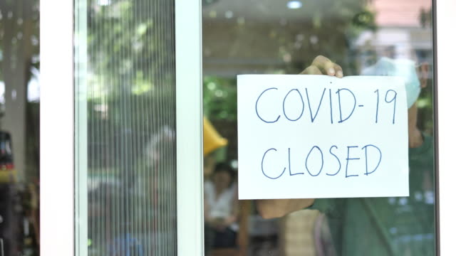 Coronavirus closed for business concept, male with medical mask puts sorry closed sign on window due to covid 19.