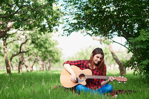 a girl in a black and red checked shirt and jeans sits on a green lawn and grass and plays a classical acoustic guitar with her own hands. A girl in nature plays a musical instrument
