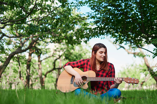 a girl in a checked shirt and jeans is sitting on a green lawn, playing a classical acoustic guitar with her own hands and singing. A girl in nature plays a musical ins