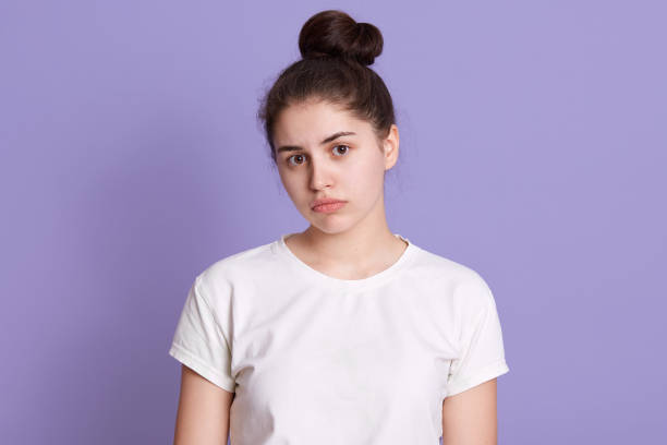 istock Dark haired lady with sad facial expression posing against lilac wall, looks at camera, being in bad mood, dresses white t shirt, stands with upset look. 1249382820