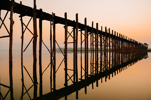 People cross the famous U Bein Bridge in the Amarapura Township of Mandalay at sunset, with reflections visible in Lake Taungthaman.