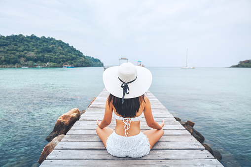 Summer travel vacations concept, Happy traveler asian woman with white bikini and hat relax and yoga meditation on wooden bridge in tropical beach, Thailand