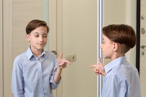 A brunette boy straightens a blue shirt in front of a mirror and shows a super sign. The child is preparing at school.