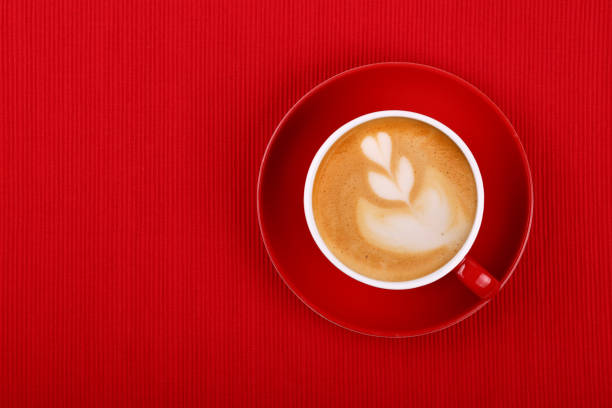 Close up one cup full of cappuccino coffee on red saucer over red tablecloth, elevated top view, directly above