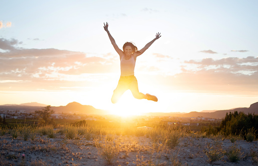 Athletic young girl jumping freely at mountain top against sunset in Murcia, Spain. Doing exercise work out, training. Attractive fit healthy woman in grass field smiling in the air. Fitness.