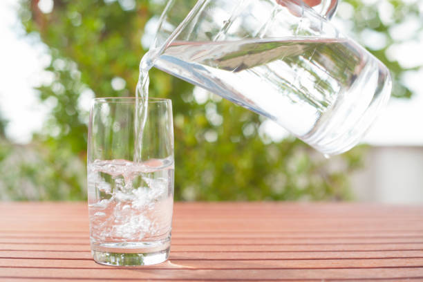 istock Drinking water flowing from a glass jug into a glass  background with plants 1249408795