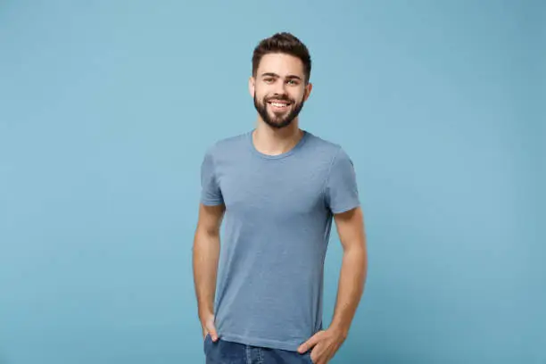 Photo of Young smiling handsome man in casual clothes posing isolated on blue wall background, studio portrait. People sincere emotions lifestyle concept. Mock up copy space. Holding hands in pockets.