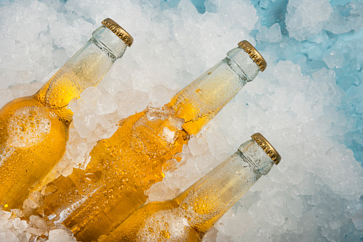 Close up three clear glass bottles of cold lager beer on crushed ice at retail display, elevated high angle view