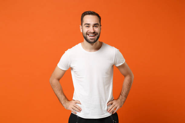 Cheerful young handsome man in casual white t-shirt posing isolated on orange wall background studio portrait. People sincere emotions lifestyle concept. Mock up copy space. Standing with arms akimbo. Cheerful young handsome man in casual white t-shirt posing isolated on orange wall background studio portrait. People sincere emotions lifestyle concept. Mock up copy space. Standing with arms akimbo isolated color stock pictures, royalty-free photos & images