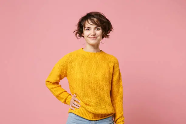 Photo of Smiling young brunette woman girl in yellow sweater posing isolated on pastel pink wall background, studio portrait. People sincere emotions lifestyle concept. Mock up copy space. Looking camera.