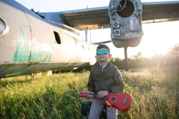 A cute boy in a bomber jacket with pilot sunglasses is sitting on a suitcase and holding a ukulele in tall grass under a wing of a decommissioned airplane in a public field in Nis, Serbia, no entry fee