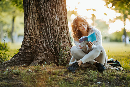 Beautiful young woman sitting under the tree in park and reading book