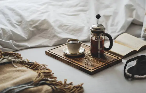 Coffee brewed in a French press and a cup on a wooden board with open book in bed. Morning at home, coziness and comfort. High quality photo