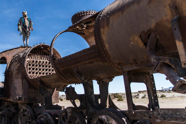 man standing on top of an abandoned train in a train cemetery with a beautiful blue sky in the background