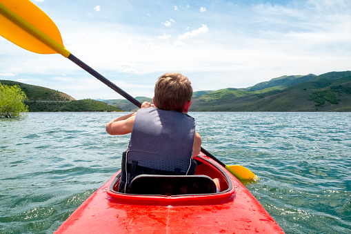 This shot was taken from the back of a two person kayak during a paddle on a mountain lake in Utah.  In front is a young boy seen from behind.  Surrounding the lake are green hills and mountains.