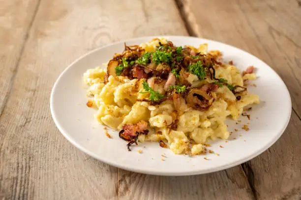 Homemade spaetzle, German egg noodles with cheese, roasted onions, bacon and breadcrumbs, served with parsley garnish on a white plate on a rustic wooden table, selected focus, narrow depth of field