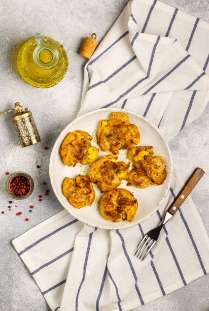 baked whole crushed and crusty potato with crust sea salt, olive oil, garlic, pepper and thyme in a white plate on a grey concrete background. roasted smashed potatoes. australian cuisine dish. selective focus - smashed potatoes imagens e fotografias de stock