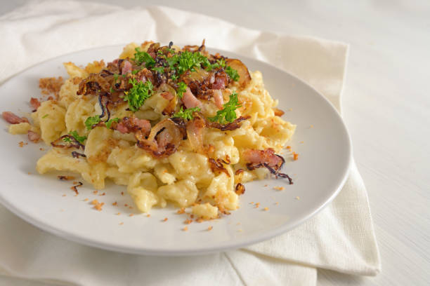 istock Homemade German egg pasta, called spaetzle, with cheese, roasted bacon, onion and breadcrumbs, served with parsley garnish on a plate, napkin and a white painted wooden table, traditional dish in southern Germany and Austria, copy space, selected focus 1249459942