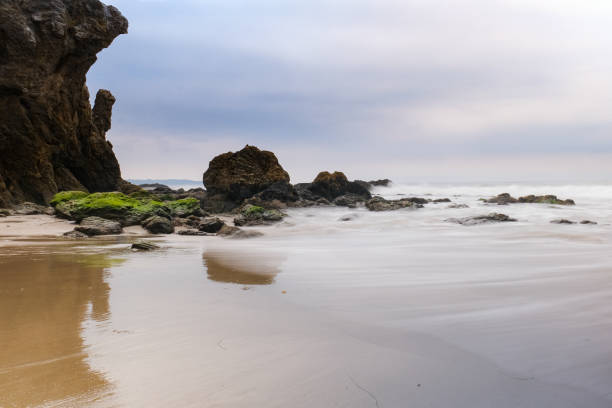 The iconic cliffs and boulders reflect in the shallow surf on Malibu's sandy El Matador Beach. The sky is soft and overcast with a thin marine layer. The waves are soft and white due to a long exposure.