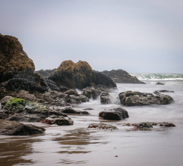 The iconic cliffs and boulders reflect in the shallow surf on Malibu's sandy El Matador Beach. The sky is soft and overcast with a thin marine layer. The waves are soft and white due to a long exposure.