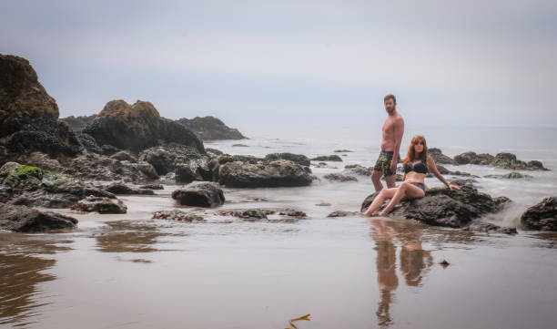 A young couple (30 something) poses together in the surf of Malibu's El Matador State Beach. They wear swim wear. The exposure is long so the water is white and soft as it moves around the feet of the models.