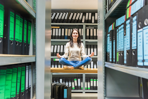 Young woman meditating with closed eyes, taking a break on office desk in between the finance file archive, surrounded with ring binders, folders, files, papers in company document storage room. Concept Shot.