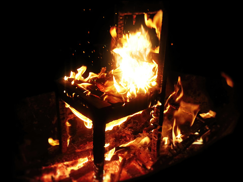 Wooden chair burning inside the bonfire. Close up on sparks rises from the fire with heatwaves at night. A horror concept of a demonic ritual or devil and demons religious worship.