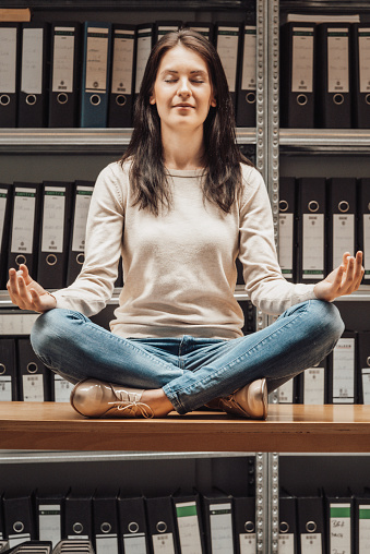 Young woman meditating with closed eyes, taking a break sitting on office desk in between the finance file archive, surrounded with ring binders, folders, files, papers in company document storage room. Concept Shot.