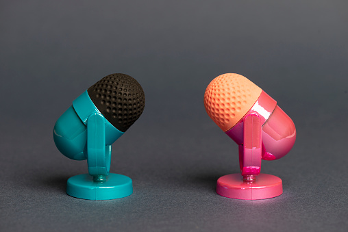 Two microphones facing each other. One microphone is pink the other microphone is blue on dark gray background. Representing a discussion between male and female arguments.