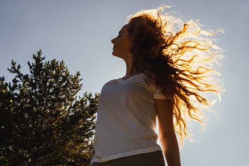 Dark silhouette of young happy girl with long curly hair in sunshine. Sky background.