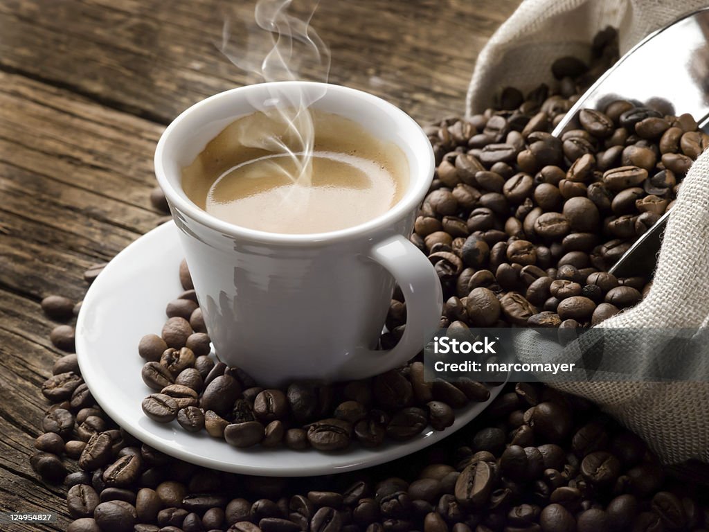 Steaming hot cup of coffee Steaming hot coffee in a small white coffee cup sits on a round saucer and is surrounded by coffee beans.  A sack of coffee beans with a shiny metal coffee scooper sticking out of it is visible along the right side of the image.  Beans are spilling from the sack onto the saucer and the brown wooden table beneath it. Espresso Stock Photo