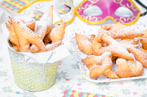 Homemade Bugnes are little golden doughnuts sprinkled with icing sugar, sweet and crispy, prepared for Mardi gras . Served in a jar with colored paper napkin, confetti and colombina carnaval masks in the background