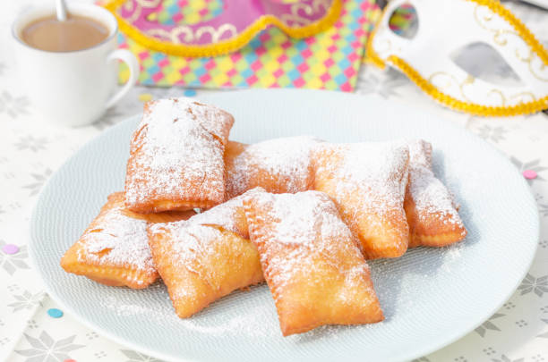 Traditional New Orleans beignets served for Mardi Gras Homemade New Orleans style beignets are small squares of fried dough covered in powdered sugar prepared for Mardi gras. Served on a plate with cup of coffee,colored paper napkin, confetti and colombina carnaval masks in the background beignet stock pictures, royalty-free photos & images