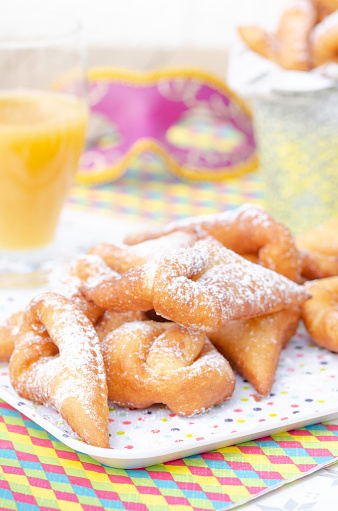 Homemade New Orleans style beignets are small squares of fried dough covered in powdered sugar prepared for Mardi gras. Served on a plate with a glass of orange juice\n,colored paper napkin, confetti and colombina carnaval mask in the background