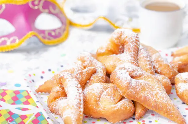 Homemade Bugnes are little golden doughnuts sprinkled with icing sugar, sweet and crispy, prepared for Mardi gras . Served on a plate with a cup of coffee, colored paper napkin, confetti and colombina carnaval masks in the background