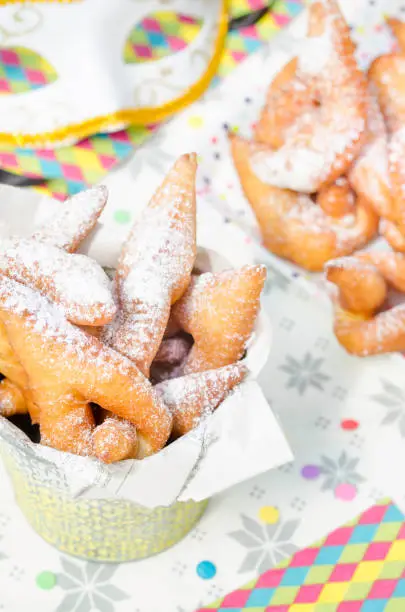 Homemade Bugnes are little golden doughnuts sprinkled with icing sugar, sweet and crispy, prepared for Mardi gras . Served in a jar with colored paper napkin, confetti and colombina carnaval mask in the background