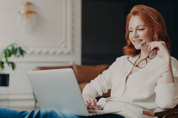 istock Pretty satisfied redhead female student watches tutorial video, works distantly from home, concentrated in laptop computer, prepares course work, holds spectacles, poses in modern spacious room 1249565488