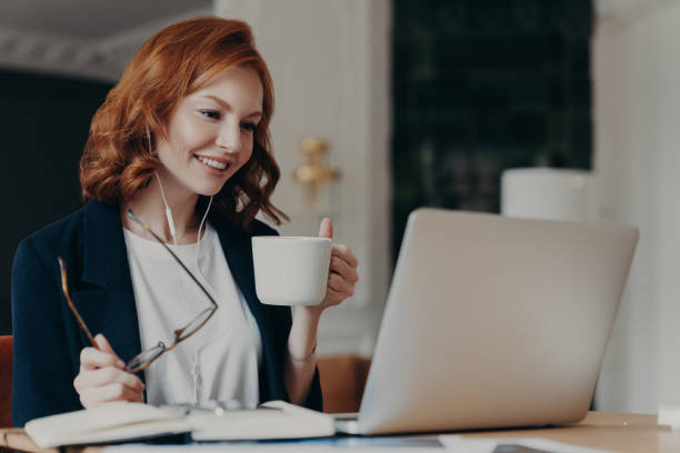 istock Intelligent female student has online course, concentrated in screen of laptop computer, learns foreign languages distantly, drinks tea or coffee, poses at desktop. Woman worker has video conference 1249565561