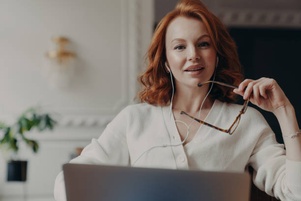 Successful redhead woman entrepreneur ready to conduct online negotiations with coworkers, holds spectacles, sits in front of opened laptop, uses earphones, searches needed information in internet exitosa pelirroja mujer empresaria lista para llevar a cabo negociaciones en línea con compañeros de trabajo, sostiene gafas, se sienta frente a la computadora portátil abierta, utiliza auriculares, busca la información necesaria en internet