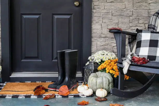 Traditional style front porch decorated for autumn with rain boots, heirloom gourds,  white pumpkins, mums and rocking chair with buffalo plaid pillow and throw blanket giving an inviting atmosphere.