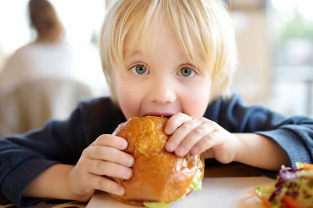 lindo chico rubio comiendo hamburguesa grande en el restaurante fastfood. comida poco saludable para los niños. comida basura. - sandwich food lunch chicken fotografías e imágenes de stock
