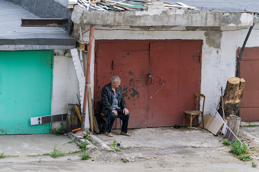 Abakan, Russia 06/14/2020:  Lonely elderly man sitting on a chair in the street near a car garage