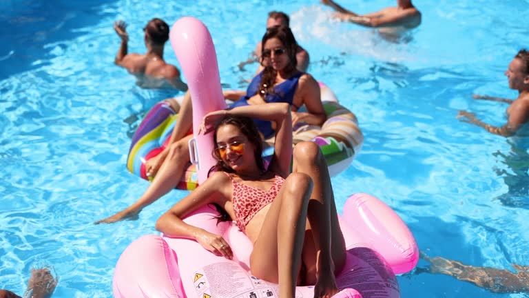 Friends chilling in private villa swimming pool, lie in the sun on inflatable flamingo, swan, floaties. Young people relax on party at luxury resort on sunny day. Bikini girls sunbathing. Slow motion