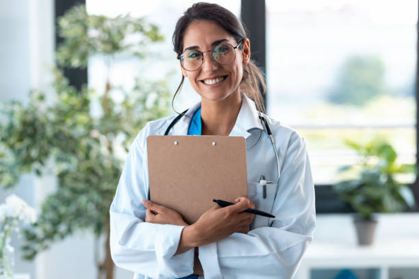 Beautiful female doctor looking at camera and smiling while holding medical documents and standing in the consultation.