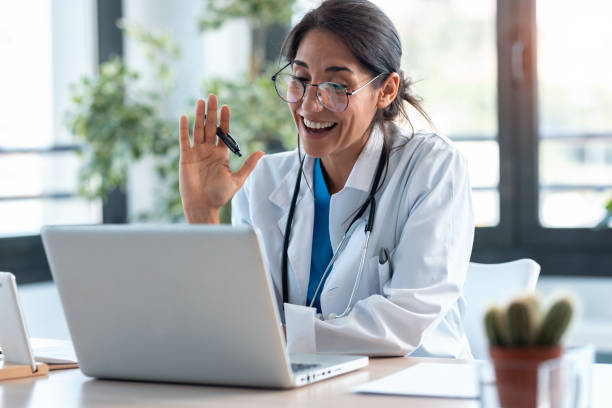 Female doctor waving and talking with colleagues through a video call with a laptop in the consultation. Shot of female doctor waving and talking with colleagues through a video call with a laptop in the consultation. telemedicine stock pictures, royalty-free photos & images