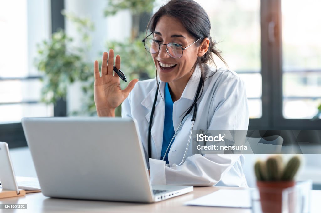 Kvinnlig läkare vinkar och pratar med kollegor genom ett videosamtal med en bärbar dator i konsultationen. - Royaltyfri Läkare Bildbanksbilder