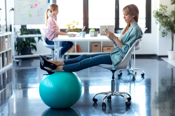 jeune femme d’affaires utilisant son téléphone portable tout en reposant des pieds sur la boule de forme physique dans le bureau. en arrière-plan, son collègue travaillant sur ordinateur portable. - yoga ball photos et images de collection