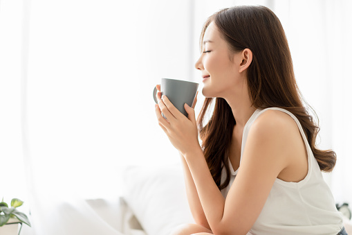 Happy smiling beautiful Asian woman enjoying first morning and smelling aroma of cup of coffee tea in her bedroom.Enjoying free and Relax time, Lazy day off concept.